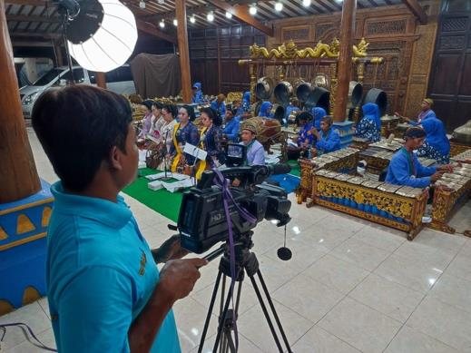 Kameramen komunitas mengoperasikan kamera di atas tripod, membidik pentas gamelan dari sisi pendopo.